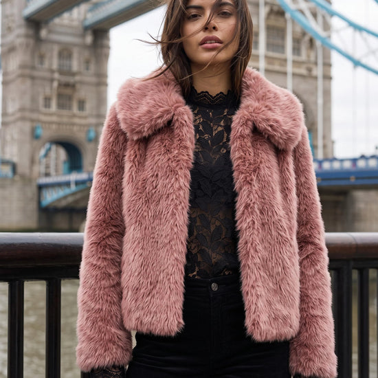 A woman wearing a pink faux fur jacket over a black lace top and black pants, standing in front of Tower Bridge in London.