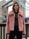 A woman wearing a pink faux fur jacket over a black lace top and black pants, standing in front of Tower Bridge in London.