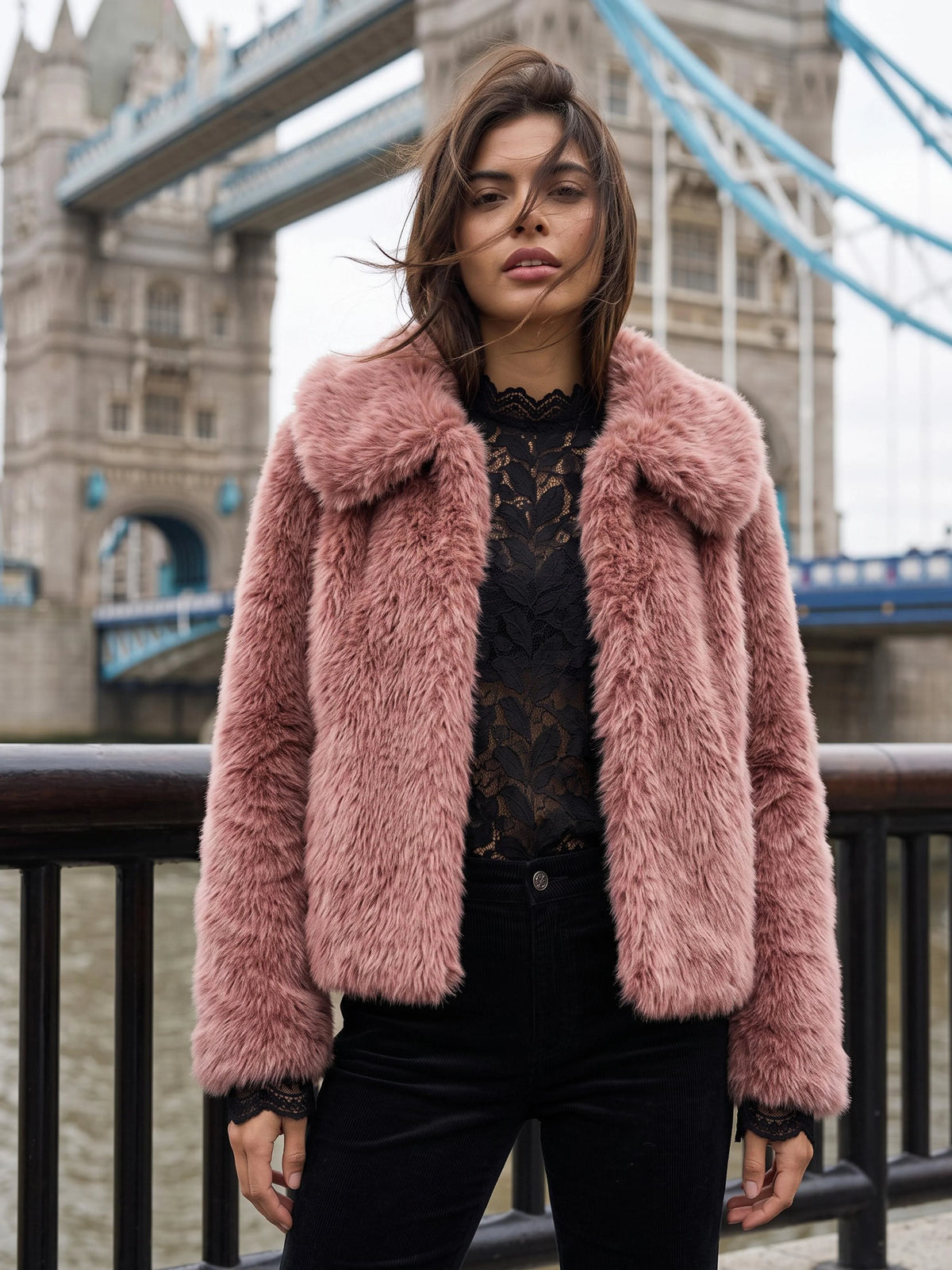 A woman wearing a pink faux fur jacket over a black lace top and black pants, standing in front of Tower Bridge in London.