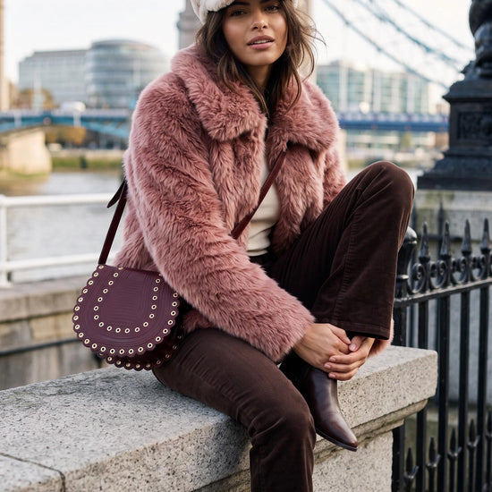A woman sits on a stone bridge railing wearing a dusty pink faux fur jacket, brown corduroy trousers, a cream beret, and a dark purple studded leather shoulder bag, with a bridge and river in the background.