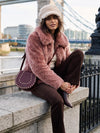 A woman sits on a stone bridge railing wearing a dusty pink faux fur jacket, brown corduroy trousers, a cream beret, and a dark purple studded leather shoulder bag, with a bridge and river in the background.