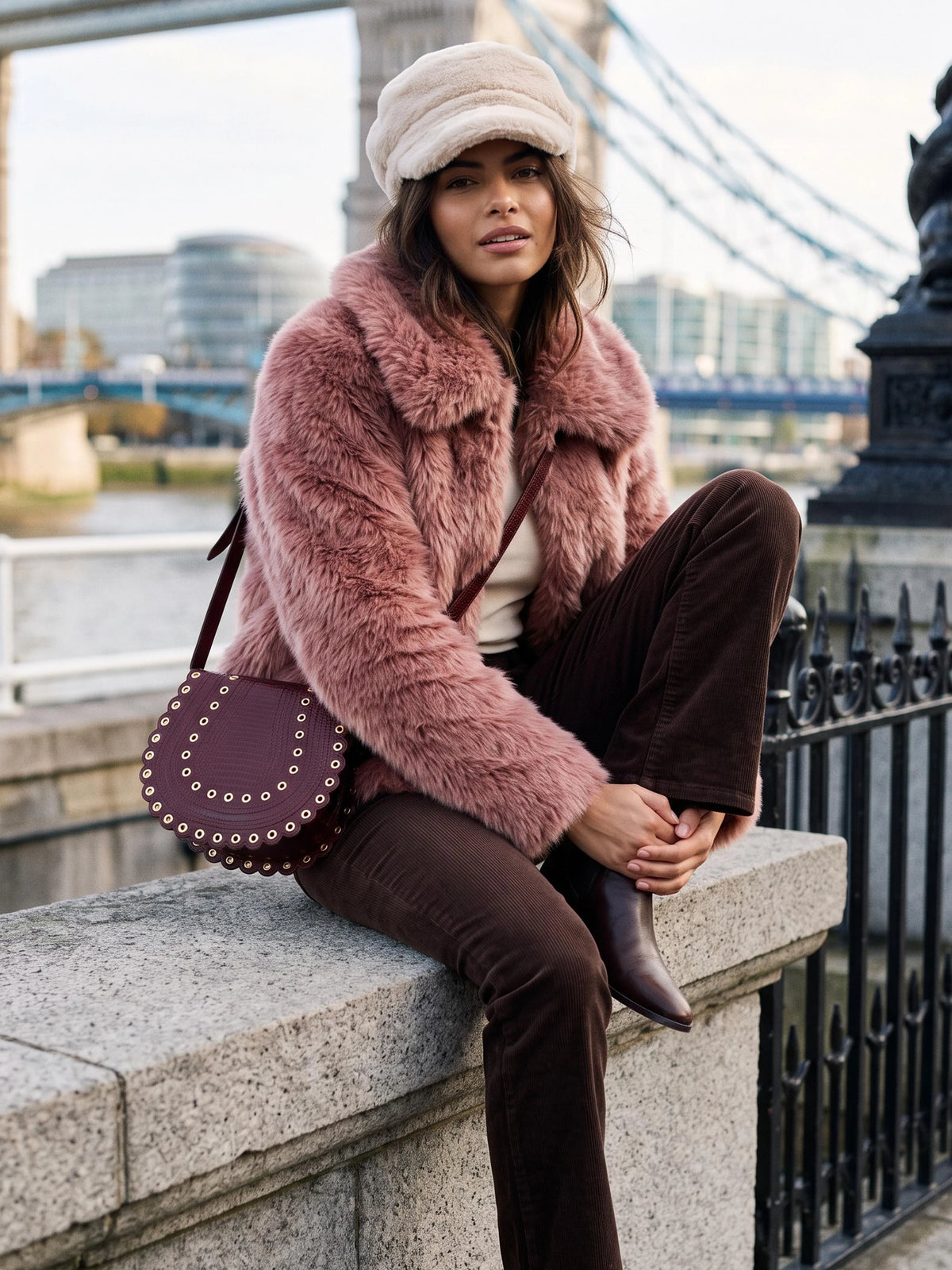 A woman sits on a stone bridge railing wearing a dusty pink faux fur jacket, brown corduroy trousers, a cream beret, and a dark purple studded leather shoulder bag, with a bridge and river in the background.