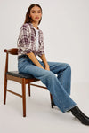A woman wearing a long-sleeve plaid button-up shirt in cream and burgundy, tucked into blue jeans with a black belt.