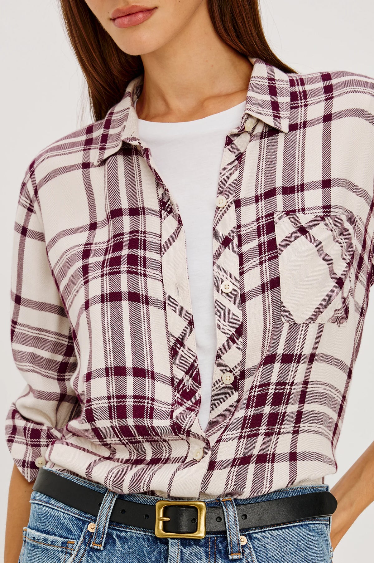 A woman wearing a long-sleeve plaid button-up shirt in cream and burgundy, tucked into blue jeans with a black belt.