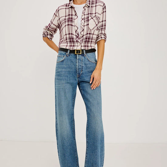A woman wearing a long-sleeve plaid button-up shirt in cream and burgundy, tucked into blue jeans with a black belt.