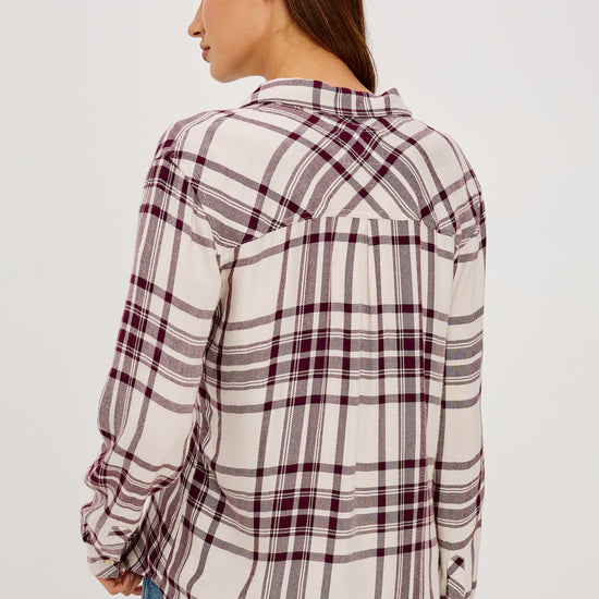 A woman wearing a long-sleeve plaid button-up shirt in cream and burgundy, tucked into blue jeans with a black belt.