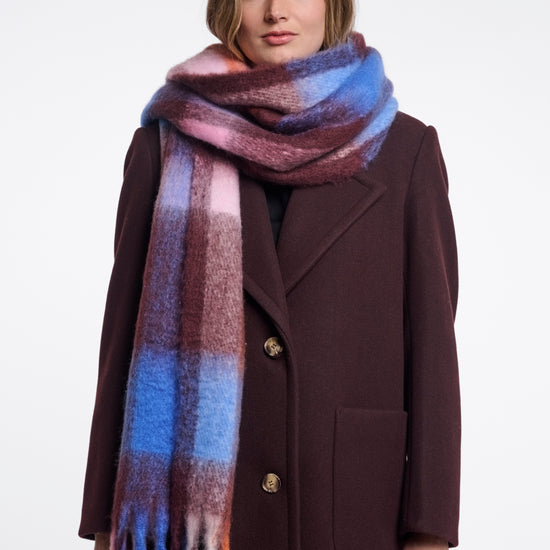 A woman wearing a dark brown wool coat and a thick, soft plaid scarf with blue, pink, purple, and orange tones, photographed against a plain white studio background.