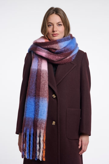 A woman wearing a dark brown wool coat and a thick, soft plaid scarf with blue, pink, purple, and orange tones, photographed against a plain white studio background.