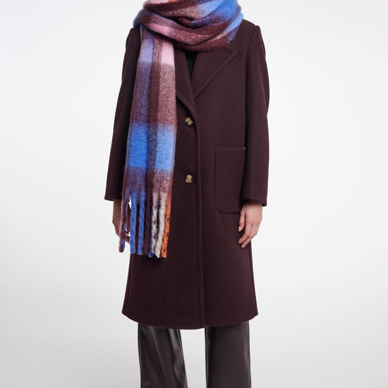A model stands against a light gray studio background wearing a long dark brown coat, wide-leg dark brown pants, and a thick plaid scarf with blue, purple, pink, and orange tones.