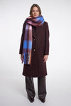 A model stands against a light gray studio background wearing a long dark brown coat, wide-leg dark brown pants, and a thick plaid scarf with blue, purple, pink, and orange tones.