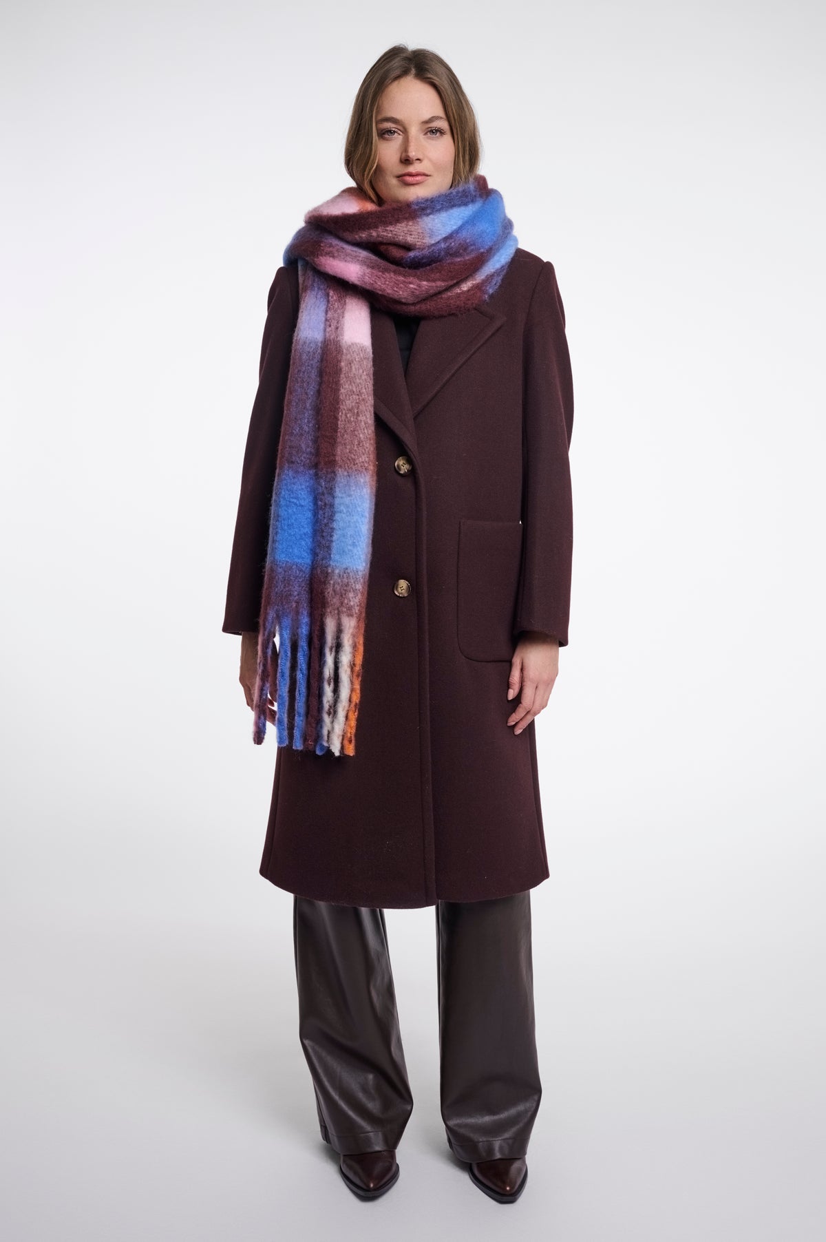 A model stands against a light gray studio background wearing a long dark brown coat, wide-leg dark brown pants, and a thick plaid scarf with blue, purple, pink, and orange tones.