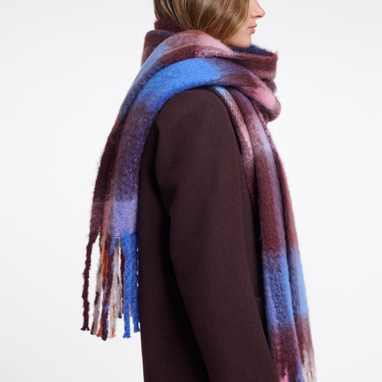 A woman shown in profile wearing a long, soft scarf with a mottled gradient of blue, purple, and burgundy tones, paired with a dark brown coat against a plain light gray background.