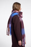 A woman shown in profile wearing a long, soft scarf with a mottled gradient of blue, purple, and burgundy tones, paired with a dark brown coat against a plain light gray background.