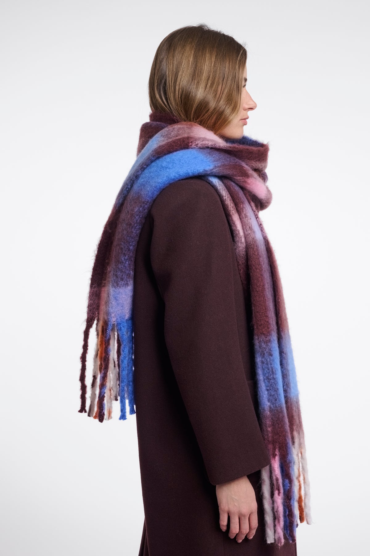 A woman shown in profile wearing a long, soft scarf with a mottled gradient of blue, purple, and burgundy tones, paired with a dark brown coat against a plain light gray background.