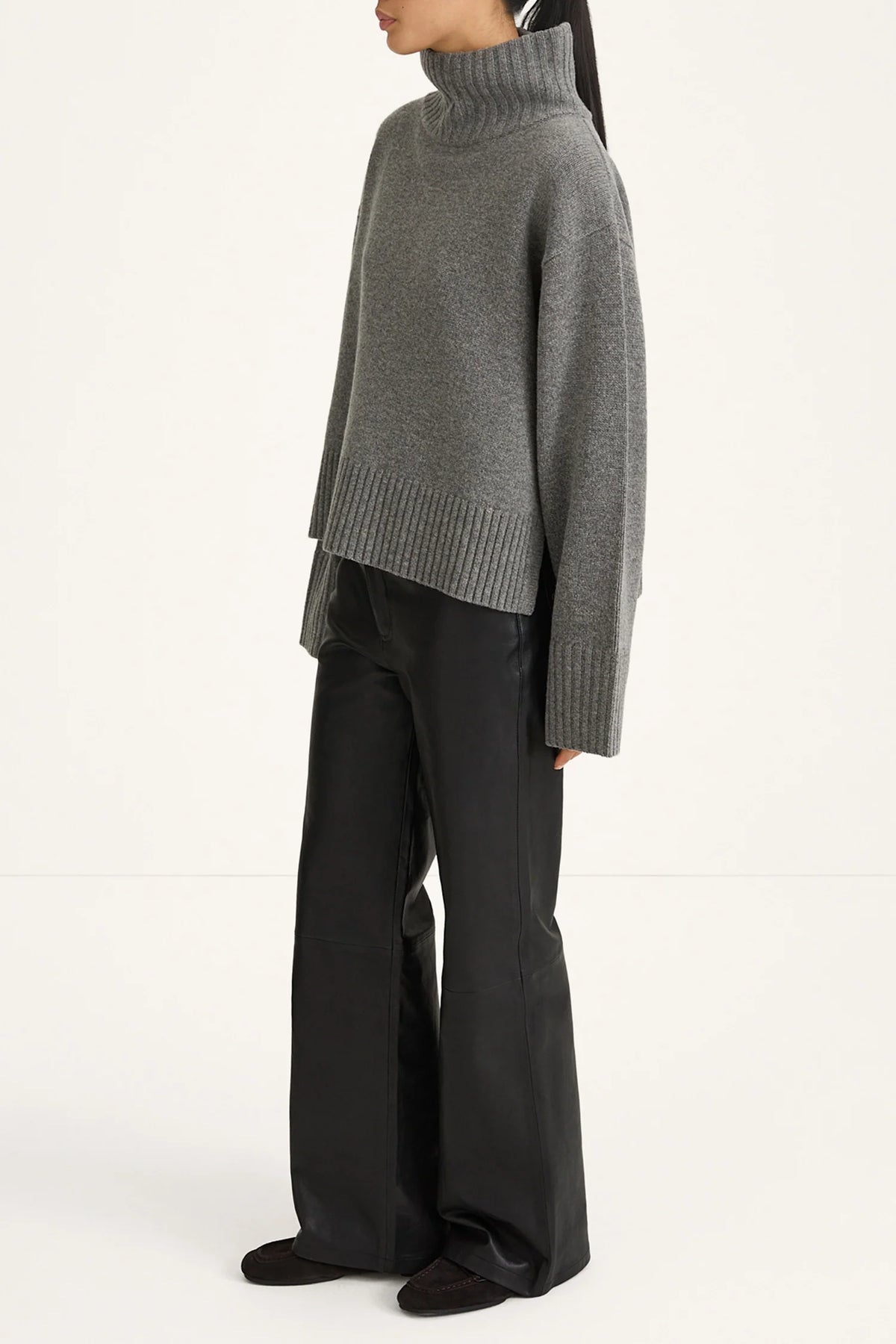 A model shown from the side wearing a gray ribbed turtleneck sweater and black wide-leg trousers against a plain white studio background.