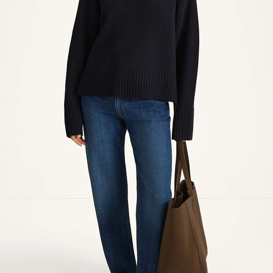 Person wearing a black turtleneck sweater, blue jeans, and holding a brown bag on a white background
