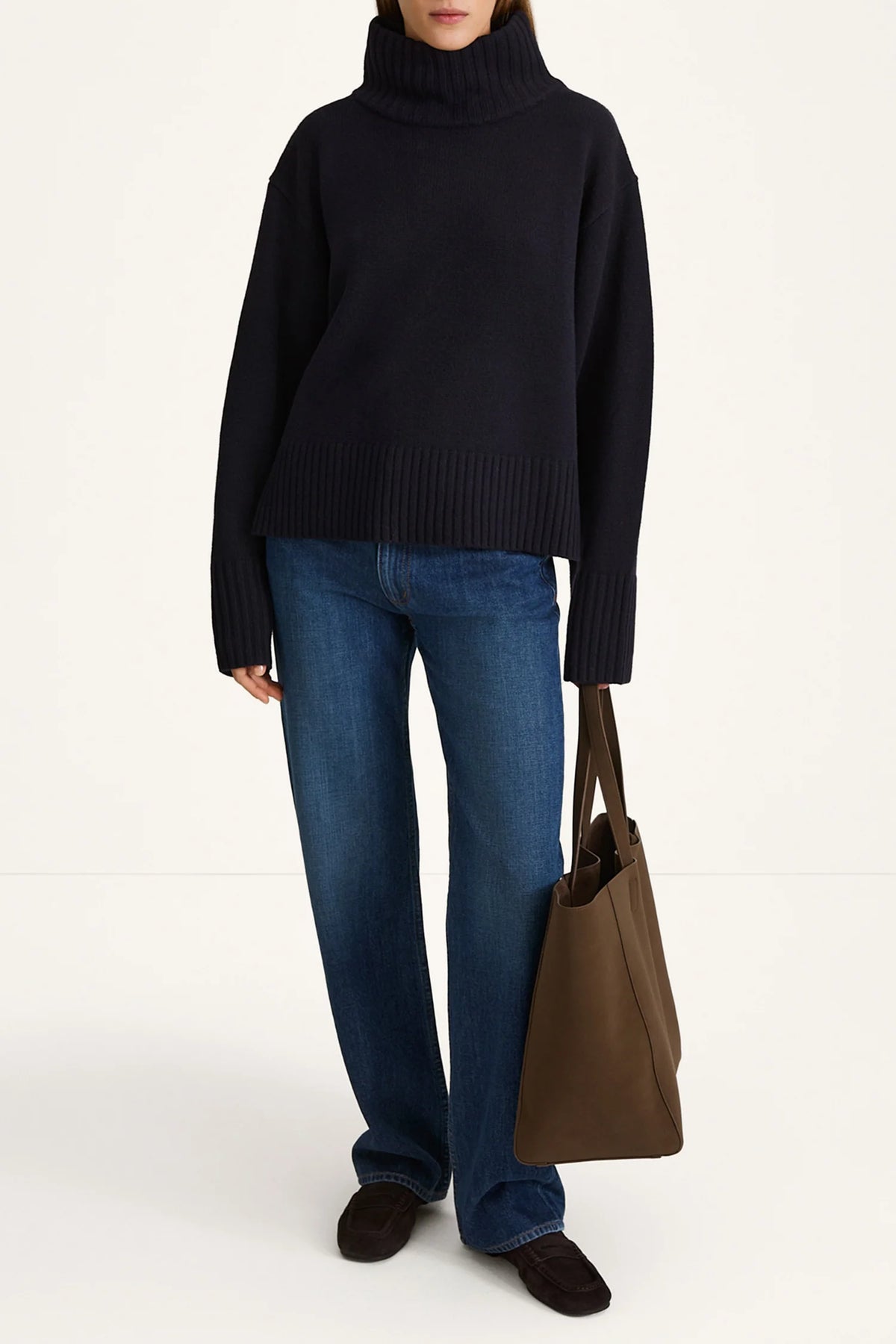 Person wearing a black turtleneck sweater, blue jeans, and holding a brown bag on a white background