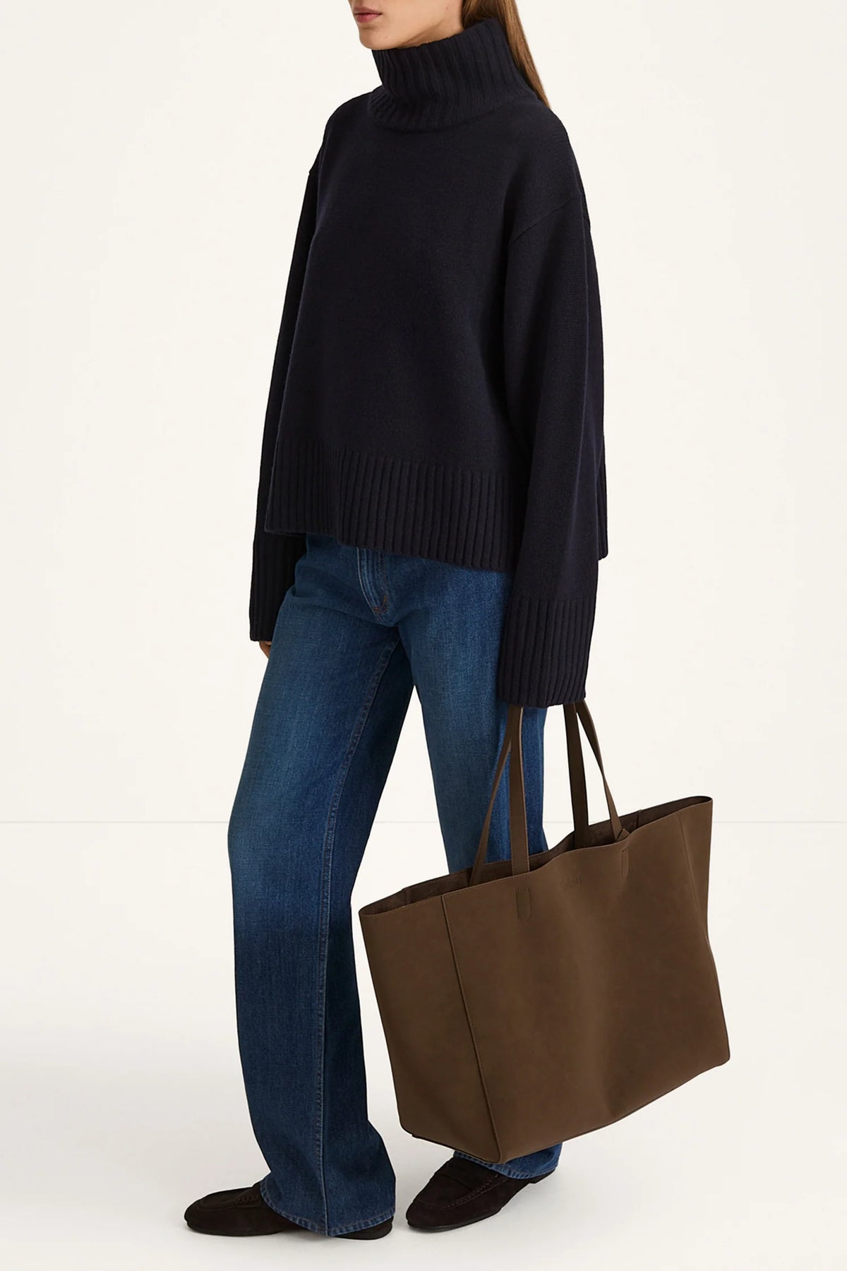 Person wearing a black sweater and blue jeans holding a brown tote bag on a white background