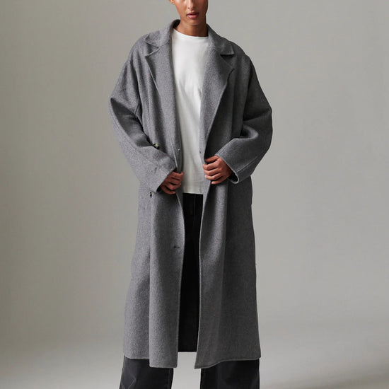 Person wearing a long gray coat over a white shirt and black pants on a plain background