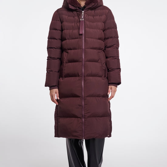 A woman modeling a long, dark plum-brown puffer coat with horizontal quilting, a high padded collar, and a front zipper, paired with black pants, against a plain light gray studio background.