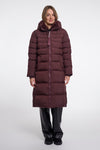 A woman modeling a long, dark plum-brown puffer coat with horizontal quilting, a high padded collar, and a front zipper, paired with black pants, against a plain light gray studio background.