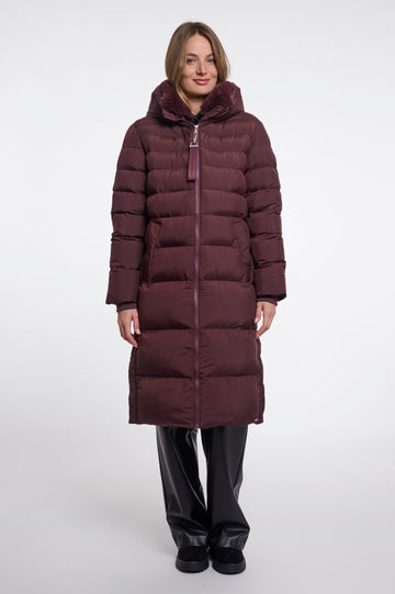 A woman modeling a long, dark plum-brown puffer coat with horizontal quilting, a high padded collar, and a front zipper, paired with black pants, against a plain light gray studio background.