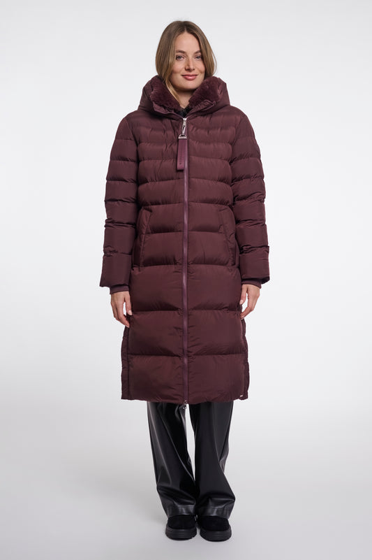 A woman modeling a long, dark plum-brown puffer coat with horizontal quilting, a high padded collar, and a front zipper, paired with black pants, against a plain light gray studio background.
