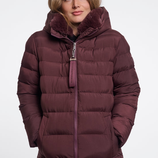 A woman wearing a long, dark plum-brown puffer coat with a plush faux-fur lined hood and a matching zipper pull, photographed against a plain light gray studio background.