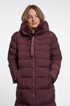 A woman wearing a long, dark plum-brown puffer coat with a plush faux-fur lined hood and a matching zipper pull, photographed against a plain light gray studio background.