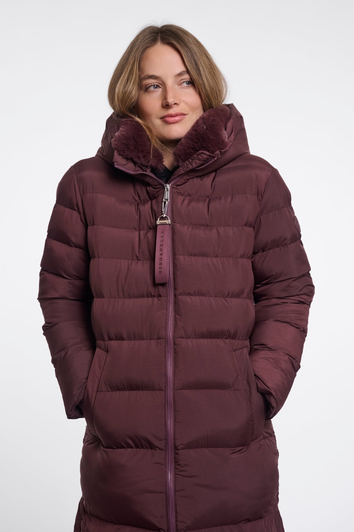 A woman wearing a long, dark plum-brown puffer coat with a plush faux-fur lined hood and a matching zipper pull, photographed against a plain light gray studio background.