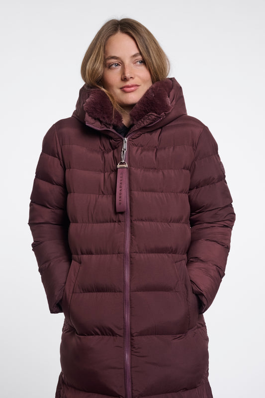 A woman wearing a long, dark plum-brown puffer coat with a plush faux-fur lined hood and a matching zipper pull, photographed against a plain light gray studio background.