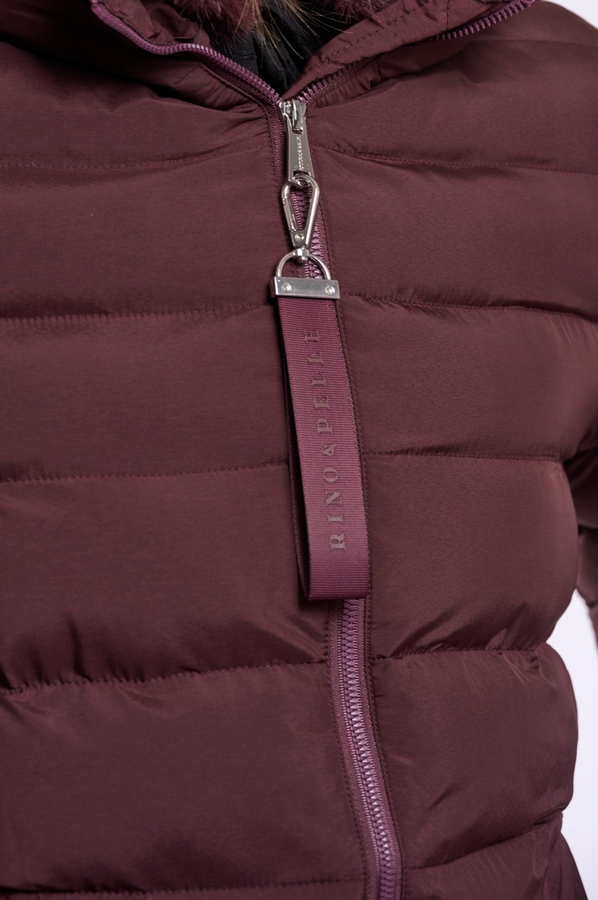Close-up of a quilted puffer jacket in a deep plum-burgundy color, featuring a silver-tone zipper and a matching fabric pull tab with the brand name 'BENDADETE' printed vertically.
