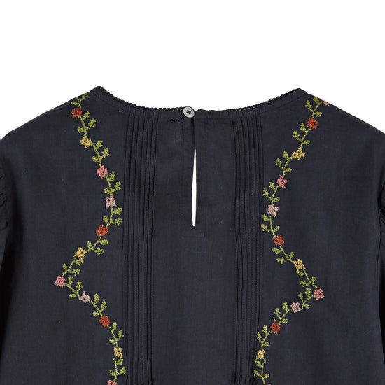 Close-up of the back neckline of a dark navy blue top, featuring vertical pleats down the center, a small button closure, and floral embroidery with green stems and pink, red, and yellow flowers along the shoulders.