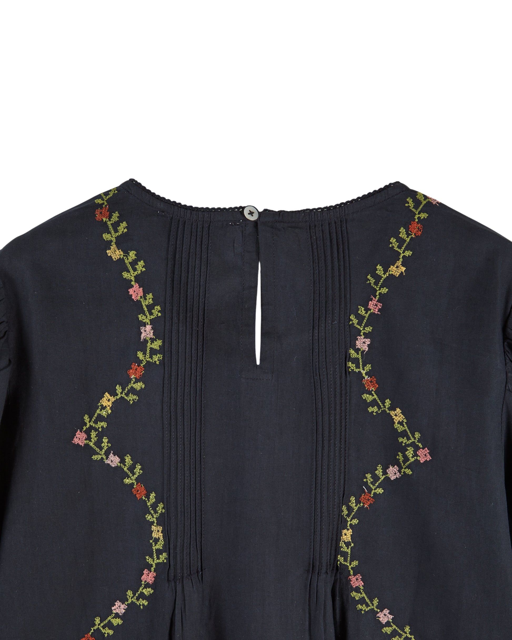 Close-up of the back neckline of a dark navy blue top, featuring vertical pleats down the center, a small button closure, and floral embroidery with green stems and pink, red, and yellow flowers along the shoulders.