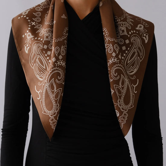A brown silk scarf with a white paisley pattern draped over the shoulders of a model wearing a black long-sleeve top, against a neutral gray background.