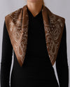 A brown silk scarf with a white paisley pattern draped over the shoulders of a model wearing a black long-sleeve top, against a neutral gray background.