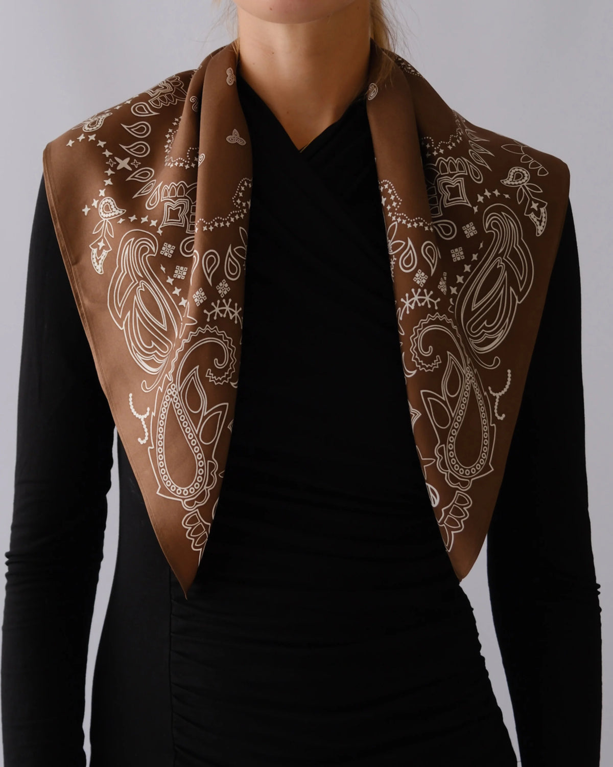 A brown silk scarf with a white paisley pattern draped over the shoulders of a model wearing a black long-sleeve top, against a neutral gray background.