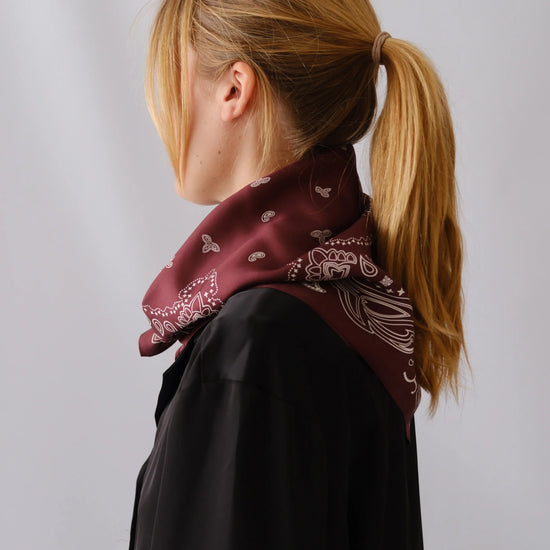 A woman with blonde hair in a ponytail is shown from the side/back wearing a dark maroon bandana scarf with a white paisley pattern over a black top, against a light gray studio background.