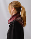 A woman with blonde hair in a ponytail is shown from the side/back wearing a dark maroon bandana scarf with a white paisley pattern over a black top, against a light gray studio background.