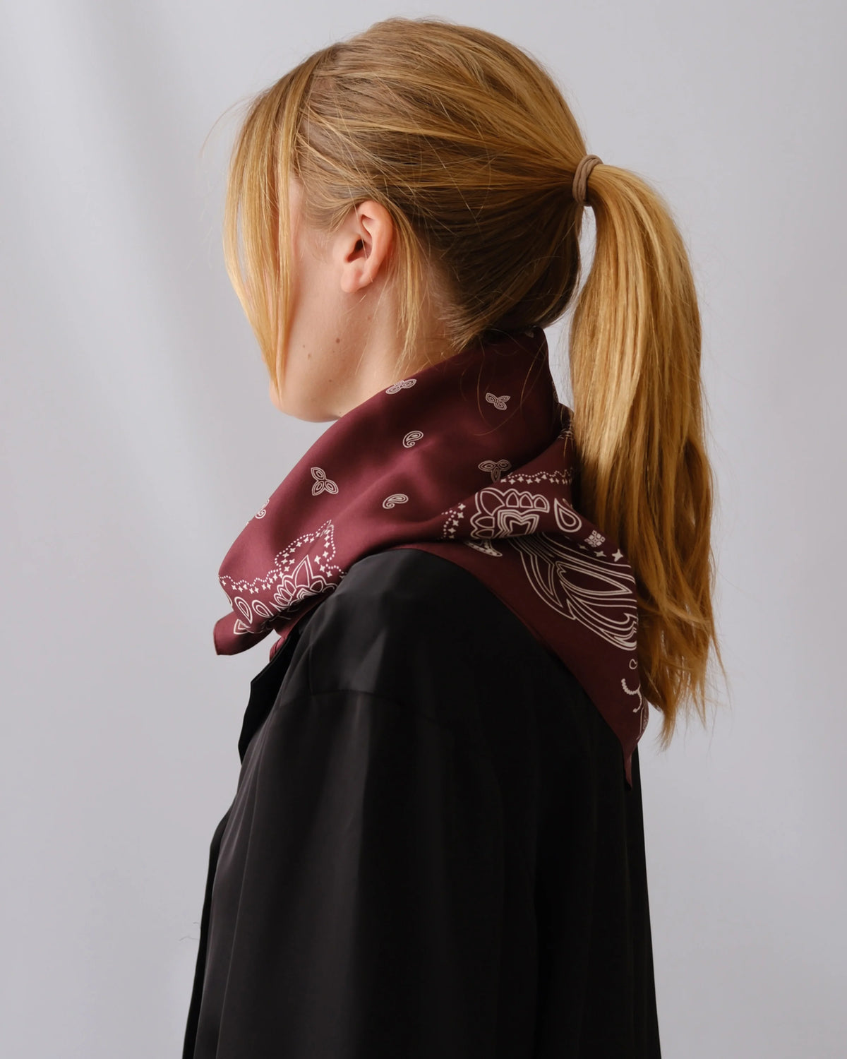 A woman with blonde hair in a ponytail is shown from the side/back wearing a dark maroon bandana scarf with a white paisley pattern over a black top, against a light gray studio background.
