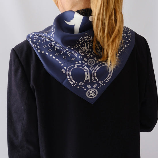 A navy blue bandana with a white western-style paisley pattern and horseshoe motif, worn around the neck of a model wearing a black top against a light gray background.