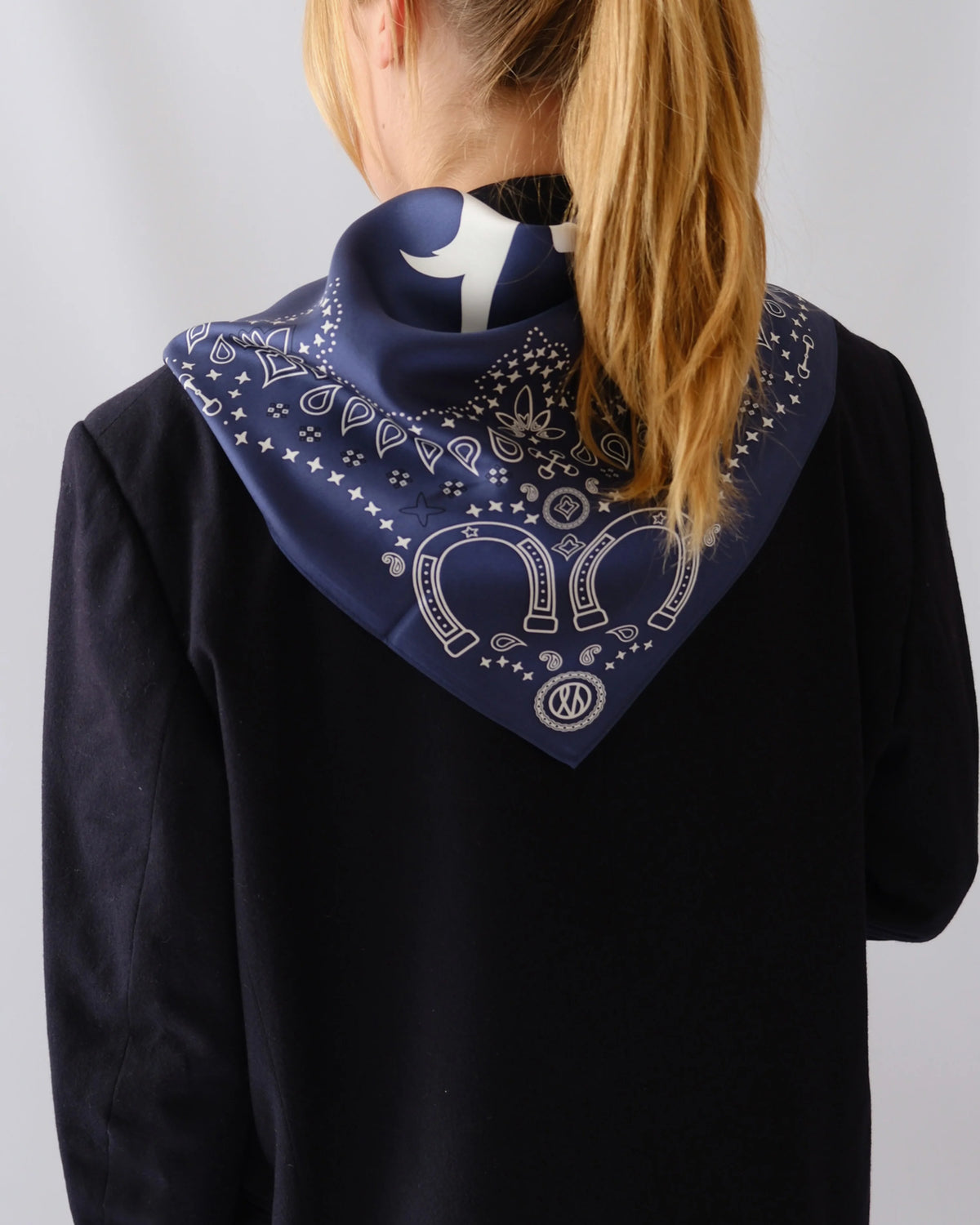A navy blue bandana with a white western-style paisley pattern and horseshoe motif, worn around the neck of a model wearing a black top against a light gray background.