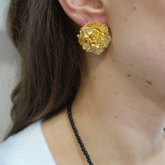 Gold flower earring worn by a person with a black beaded necklace.