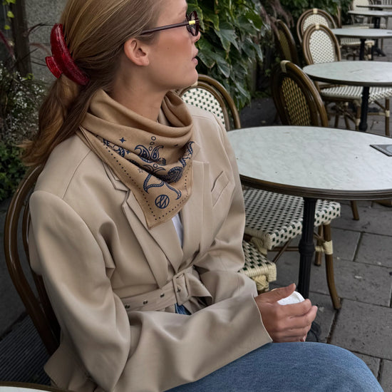 Woman sitting outdoors at a cafe table wearing a beige coat and a lescarf wool and silk blend