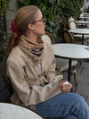Woman sitting outdoors at a cafe table wearing a beige coat and a lescarf wool and silk blend