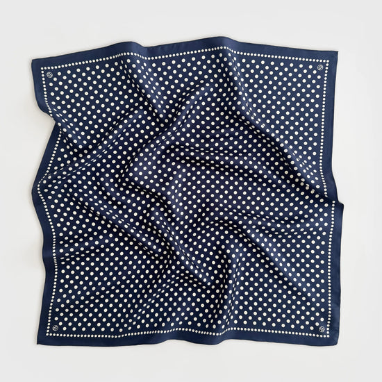A square navy blue silk scarf with a white polka dot pattern and a dotted border, laid flat on a light gray background.