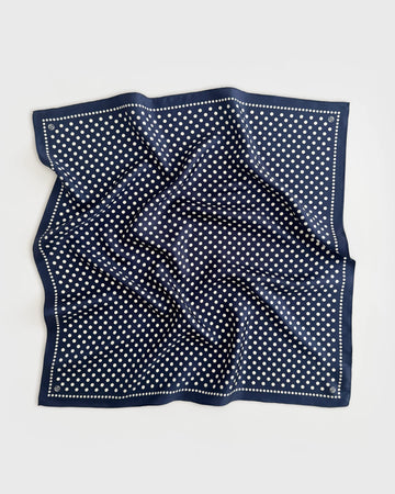 A square navy blue silk scarf with a white polka dot pattern and a dotted border, laid flat on a light gray background.