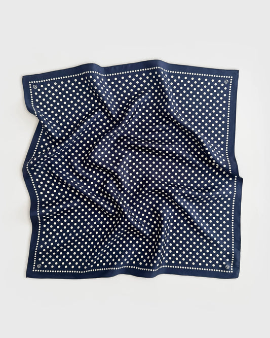 A square navy blue silk scarf with a white polka dot pattern and a dotted border, laid flat on a light gray background.