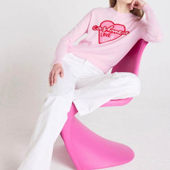 Person wearing a pink sweatshirt with text and white pants sitting on a pink chair against a light gray background
