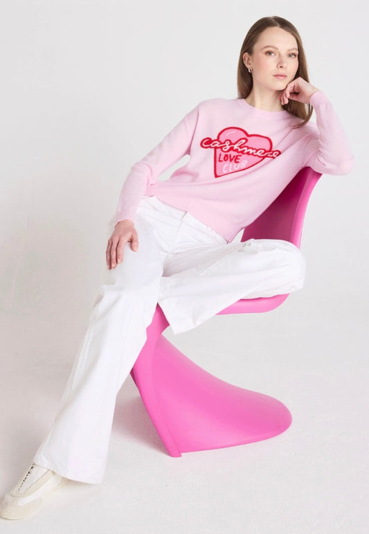 Person wearing a pink sweatshirt with text and white pants sitting on a pink chair against a light gray background